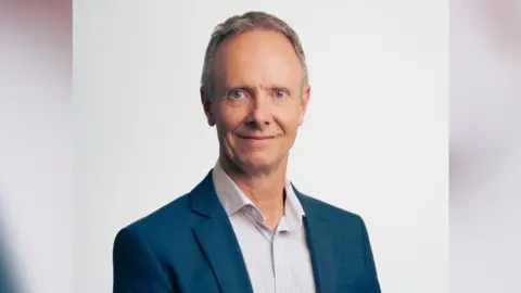 RAC Headshot of Simon Williams. He wears a navy jacket and a light pink and white shirt with no tie. The background of the image is white. 