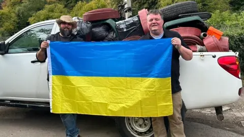 Councillor John Lawson and another man holding up a Ukrainian flag in front of a white pick up truck full of emergency aid supplies 