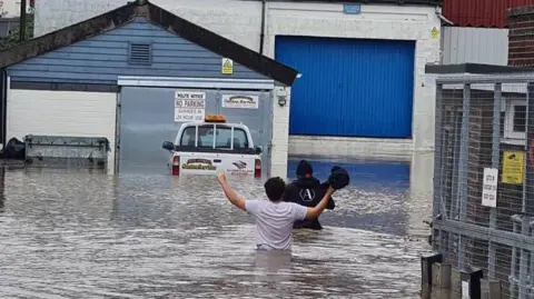 Two people walk through waist-deep floodwater outside commercial premises behind a wire fence.