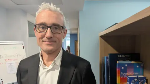Ian is standing indoors beside a tall wooden bookcase filled with academic‑style books. Several large hardback volumes are visible on the shelf, including titles related to psychology. He is wearing a dark blazer over a light‑coloured button‑up shirt. To the left, a whiteboard on a stand contains handwritten notes