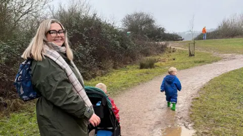 Evie Brooks A woman with blonde hair and black glasses smiling at the camera. She is wearing a green jacket and a scarf. She is outside in a park with her two sons, one of which is in view and is wearing a blue raincoat and green wellies. 