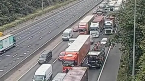 Queuing traffic on the M25 . There are a mix of lorries, vans and cars tightly packed across three lanes of traffic on the road.