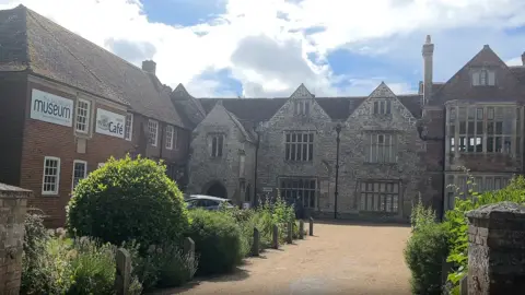 View on a sunny day of The Salisbury Museum. There is a very old grey stone building directly alongside a more modern-looking red brick building. There is a gravel driveway which leads up to the front of the museum with shrubbery on either side of it.