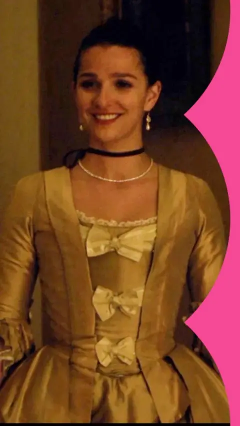 A woman smiling in a 1700s style golden dress with three bows on the front