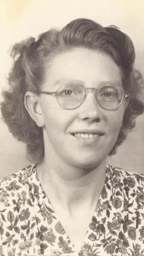 Ingrid Lyon A sepia-toned photo of Mary Meijer. She is wearing spectacles and a floral top. The front and side sections of her short hair is pinned back and she is smiling at the camera.