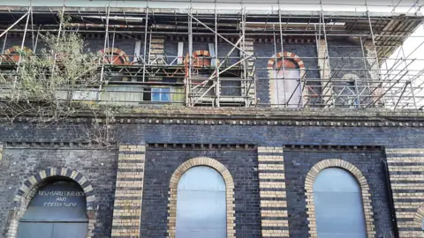 Victorian Society The black and beige building can be seen with scaffolding around the top part. A lower level of black bricks and an arched pattern of beige bricks remains untouched.