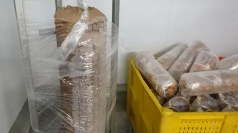 Roll of kebab meat on large spit wrapped in plastic with more rolls packed and in a yellow crate to the right