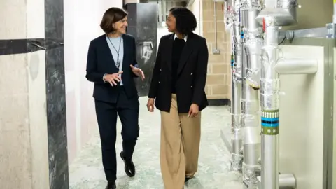 Robert Thom / Ministry of Justice. Two smiling women walk through a building with silver pipe work to the right of them. It appears that work is still ongoing.