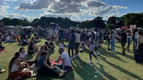Bedford Borough Council A large group of people at a festival sitting or standing on a grassy field. Stalls and trees surround the edge.