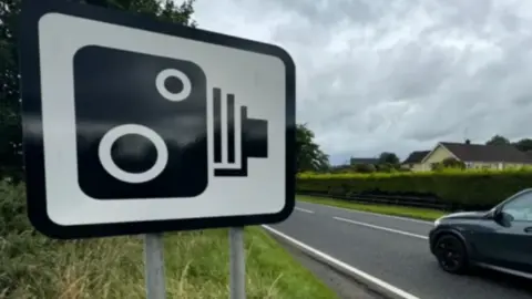 A stock image of a speed camera sign beside a B road. A car can be seen driving past the sign.