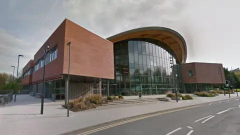 A large, glass-front building at the University of Warwick. The pavement and road in front of the building are also visible, and bushes line the steps and entrances to the building.