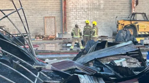 Fire fighters stand in a scrap metal yard with large sheets of metal lying around in piles. 