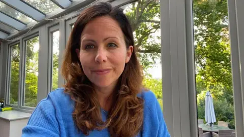 Beth Fordham, a woman with long brown hair and a blue top, is sitting in a conservatory.
