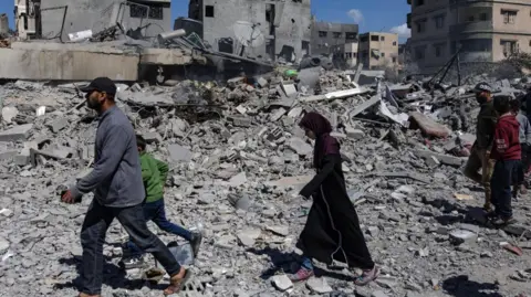 Two men, three boys and one girl walking across the rubble following an Israeli airstrike on Gaza on Wednesday, with some buildings in the background still standing.