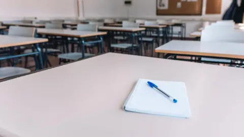 A stock picture of an empty classroom, with a notepad and blue biro on top of it.