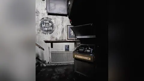 Claire Hannah Image shows fire damage to the inside of a pub. A retro jukebox is on the right with the wall, radiator and carpet showing smoke damage. 