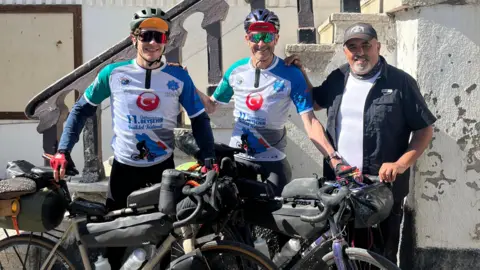 Geoff Sherwood The cyclists in special white and blue cycling vests with a turkish flag symbol on them they are stood with their bikes, wearing sunglasses and stood next to a man who is wearing a hat