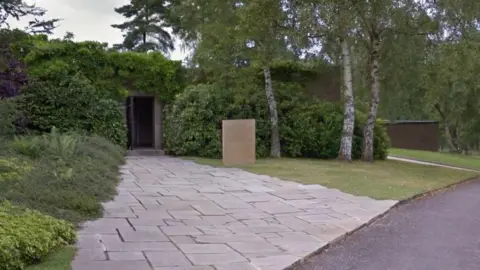 LDRS A grey, slabbed footpath that leads to a small door. Grass and trees to the right of the path, with hedges surrounding the entrance. There is a brown stone sign on the grass. 