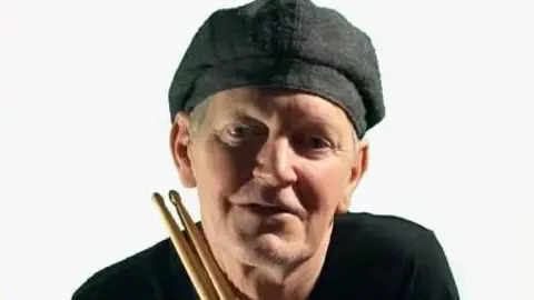 John Bennetts John Bennetts holding drumsticks and wearing a black t-shirt and grey hat, looking at the camera