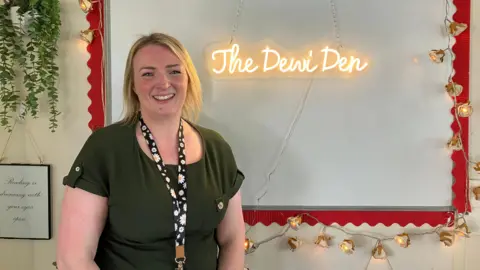 A woman with neck-length blonde hair, wearing a dark green top and a black lanyard with white daisies on it is smiling at the camera. She is standing in front of a whiteboard surrounded by lights and red trim on which an electric light reads: "The Dewi Den."