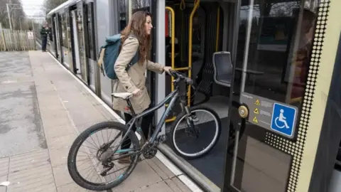 Woman with long brown hair wearing a beige coat pushes her bicycle on to a Metrolink tram