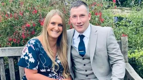 Stephanie and her late husband Martin sit on a bench in a garden on a summer day. On the left Stephanie has long blond hair and wears a navy blue dress with a floral pattern. On the right Martin wears a grey three piece suit with a navy blue tie. 