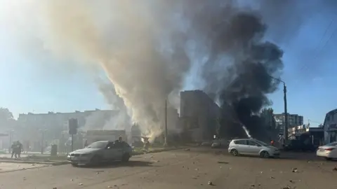 Vadym Filashkin Thick black and white smoke rises from buildings hit by a missile strike at Dobropillia, in Ukraine's eastern Donetsk region. Debris can be seen on the road with three cars pictured in the foreground.