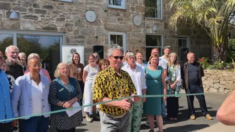 The Mayor of Penzance cuts a ribbon in front of a new building. A crowd of people behind him who were involved in the restoration project cheer. The building has old granite stonework but modern windows.