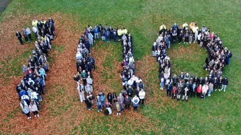 Stoke-on-Trent City Council An aerial view of people gathered together to display the number 100. They are standing on grass, which is partially covered in brown leaves.