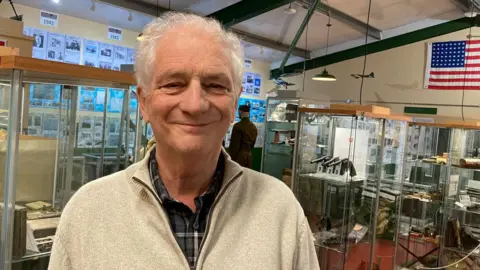 Katy Prickett/BBC Chris Pratt who has short white hair, is wearing cream half-zipped jumper over a plaid shirt and is smiling. He is standing in the British Resistance Museum, a vaulted room with glass cabinets full of decomissioned guns and rifles, wirelesses and other artefacts. 