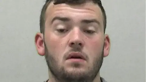 Mugshot of a man with a short whispy beard and short dark hair. He has a glazed expression on his face