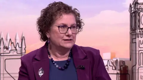 Hilary Cass on the Sunday With Laura Kuenssberg set. She has medium-length curly brown hair and is wearing a purple jacket, dark blue top and has a large purple bead necklace and purple bead earrings.