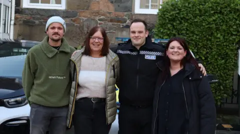 A picture of four people stood in front of a police car. There is 3 members of the public and one police officer in uniform.