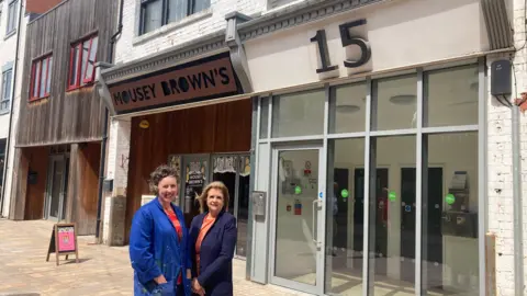BBC / Sally Fairfax Lady smiling with short wavy hair, blue denim jacket and striped red and orange jumper with lady with long brown hair, blue jacket and orange top.
They are stood on a paved street in front of wood and glass buildings with an advertising board outside.