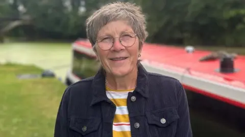 Close up of Amanda Taylor-Beswick standing outdoors beside a canal boat with a bright red roof. She has short grey hair, round glasses, and is wearing a dark jacket over a striped top. The background is softly blurred, with green grass, trees, and the canal boat stretching behind them. 