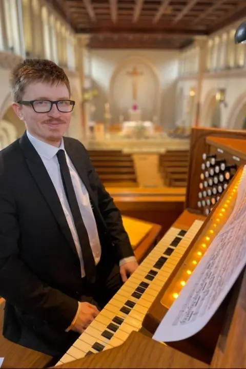 Jobe Sullivan A man with auburn hair and black glasses smiles at the camera while sitting at an organ. He is wearing a black suit and tie and a white shirt. The background of the image, which has been blurred to focus on the man, is that of a large white church with brown palaces.