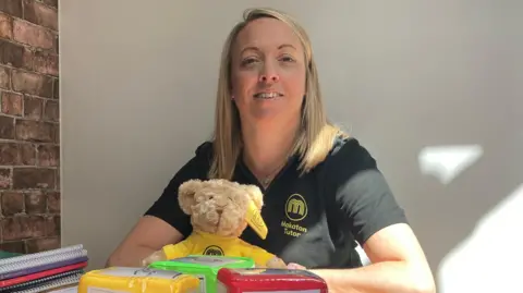 A woman with shoulder-length blonde hair is sat at a table which is covered in children's book and games. The woman is wearing a black T-short and smiles at the camera.
