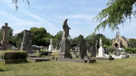 A cemetery with a variety of tombstones, stone crosses, and statues, including religious figures. In the background stands a small chapel or church with a pointed roof, surrounded by trees under a clear sky.