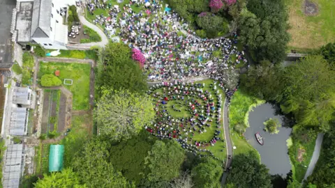 Duncan Scobie A drone shot above Helston's Flora Day shows spirals of dancing couples and a crowd watching them near a boating lake in the park.