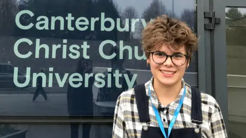 Freya Hodge - wearing a chequered shirt, a lanyard and dungarees - stands in front of a sign for Canterbury Christ Church University. 