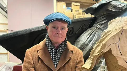 Keith Barratt is a man in his 50s wearing a brown coat with a black and white scarf and a flat cap. He is in a studio and there is part of a large sculpture just visible behind him 
