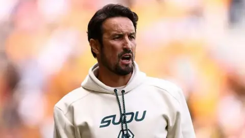 Getty Images Cesar is wearing a white hooded top and shouting directions at a player during game. He has a tan, dark hair and beard.