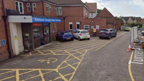 A street-view of Exmouth Hospital main entrance. There are yellow disabled parking bays outside of the entrance with cars parked on the other side. The sign at the top of the entrance is blue.  