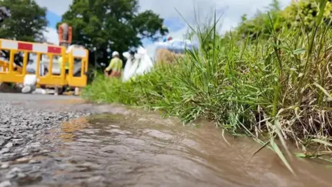 An image of people working in the road. they are fixing a leak in hard hats and high-viz. They are working behind protective fencing and there is water running down the road