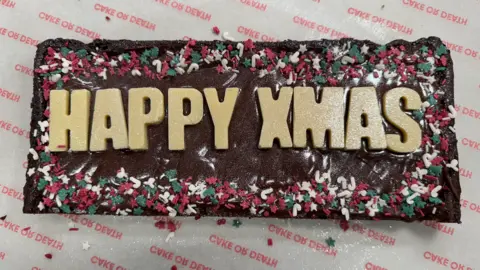 A chocolate brownie decorated for Christmas - it has the words Happy Xmas on it, brown icing and red, green and white sprinkles around the edges