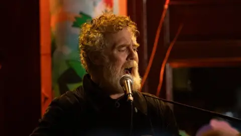 Glen Hansard has curly reddish/grey hair and beard and is singing while playing a guitar