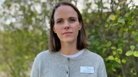 Fiona Hatton has brown straight hair that sits just above her shoulders. She has blue eyes and is wearing a grey buttoned cardigan. She is standing outside in front of leafy trees.