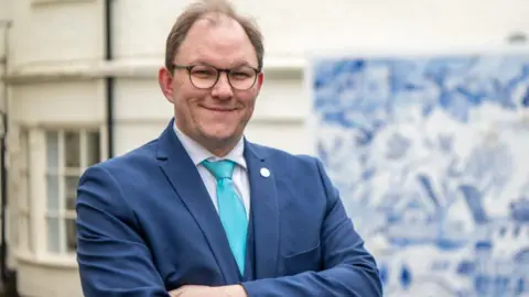 Gareth Snell Gareth Snell has his arms folded and is smiling at the camera. He is wearing a navy clue suit and light blue coloured tie. 