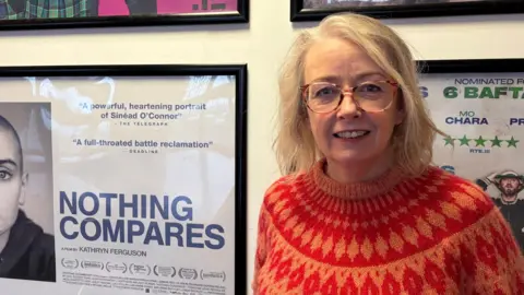 Brónagh McAtasney has shoulder-length blonde hair and is wearing glasses and a Fair Isle-style jumper in orange and red. To her right on the wall behind her is a posted advertising a film. It has a white background with large blue font reading "Nothing Compares" and half of a photograph of Sinead O'Connor is to the far side of it.