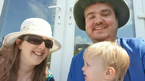 Supplied A young family play together with mum and dad in sunhats holding up a small blonde boy